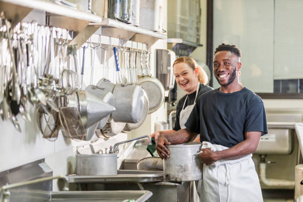 Two multi-ethnic workers in a commercial kitchen. The young African-American man is in his 20s, and the woman is in her 30s. They are both smiling and the woman is looking at the camera.